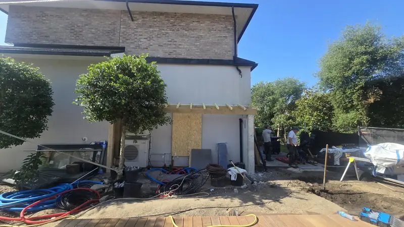 Structure en bois posée contre la façade en briques d'une maison, deux ouvriers sur le terrain